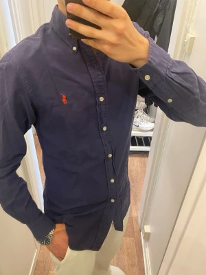 Mörkblå Skjorta från Ralph Lauren (Slim fit) - Klassisk mörkblå skjorta från Ralph Lauren med röd broderad logga på bröstet. Skjortan har knappar hela vägen, button-down krage och långa ärmar. Tillverkad i bomull för en bekväm känsla och snygg passform. Storlek Medium men passar även small. Snabb leverans!