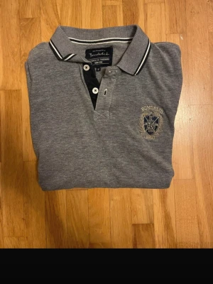 Grå pikétröja från Bondelid - Klassisk grå pikétröja från Bondelid med vit och svart rand på kragen och broderad logga på bröstet. Tröjan har knappar vid halsen och är tillverkad i mjuk bomull. Perfekt till en avslappnad men snygg stil.