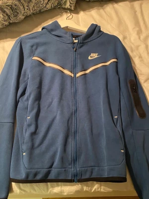 Blå Nike Tech Fleece - Blå Nike Tech Fleece med hel dragkedja och vit logga på bröstet. Har huva, vita detaljer över bröstet och en praktisk ficka med dragkedja på ärmen. Perfekt för en sportig och avslappnad stil.