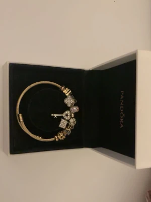 Guldigt Pandora armband med berlocker - Snyggt guldigt armband från Pandora med flera glittrande berlocker, bland annat hjärtan och nyckel. Armbandet har en elegant och modern design, perfekt för att lyfta din stil med lite extra bling.