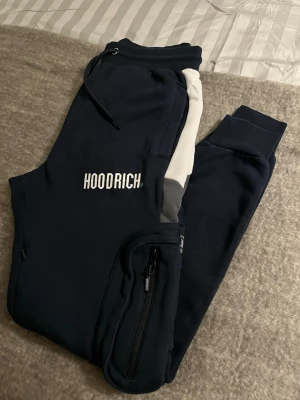 Marinblå mjukisbyxor från Hoodrich - Mörkblå mjukisbyxor från Hoodrich med vita och grå detaljer längs sidorna. Byxorna har resår i midjan med dragsko, två stora fickor med dragkedja på benen och tryckt logga på framsidan. Perfekta för en chill och sportig stil.