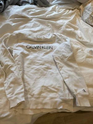 Vit sweatshirt från Calvin Klein - Snygg vit sweatshirt från Calvin Klein med svart logga framtill och liten CK-detalj på ärmen. Tröjan har rund halsringning, ribbade muddar och en patch med 'JEANS' bak. 