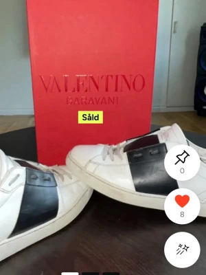 Valentino Garavani vita sneakers - Stilrena vita sneakers från Valentino Garavani med bred svart läderdetalj på sidan. Klassisk låg modell med vita snören och diskret logga. Perfekta för dig som vill ha en clean men lyxig look.