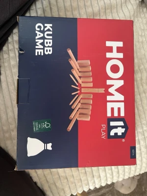 Kubbspel i trä från Home It - Säljer ett komplett kubbspel från Home It. Spelet innehåller kung, kubbar, kastpinnar och markeringspinnar, allt tillverkat i trä. Perfekt för utomhusaktiviteter med vänner eller familj. Förpackningen är blå och röd med tydlig märkning.