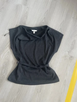 Svart topp med bälte från H&M - Grå/svart topp från H&M med bälte i midjan. Toppen har en lös passform och är ärmlös, andvönd 2 gånger, en liten fläck på baksidan, kanppt synlig, storlek S passar M också!