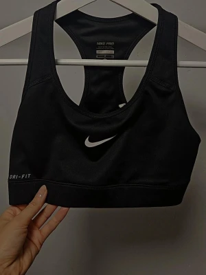 Svart sport-bh från Nike Pro - Svart sport-bh från Nike Pro med Dri-FIT-teknologi. Klassisk racerback-modell och vit Nike-logga framtill. Tillverkad i ett stretchigt och fukttransporterande material som passar perfekt för träning. Säljer för min rygg är för bred men den är helt ny.