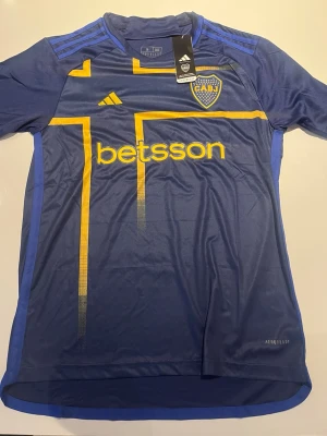 Boca Juniors matchtröja blå/gul - Snygg Boca Juniors matchtröja i blått med gula detaljer och tryck. Tröjan har klubbmärke på bröstet, sponsorloggor och är tillverkad i lätt och ventilerande material med Aeroready-teknologi. Perfekt för fotbollsträning eller att bära på läktaren.