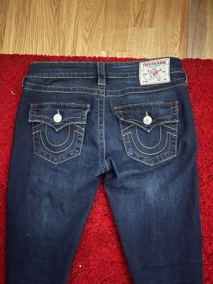 Mörkblå True Religion jeans - Snygga mörkblå jeans från True Religion med klassiska kontrastsömmar och detaljerade fickor. Modellen har låg midja och raka ben. Perfekta för dig som gillar streetwear och vill sticka ut med ett ikoniskt märke.