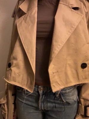 ZARA cropped trench coat - Stilren beige trenchcoat i cropped modell med breda slag, stora svarta knappar och bälte i midjan. Ifall du har XS kommer den passa!! eftersom jag har XS men jackan är i storlek S. Köptes i somras, använd 2-3 gånger( öppen för prisförslag)
