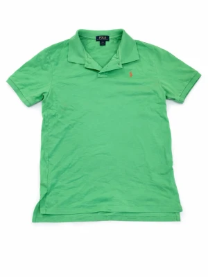 Grön pikétröja från Polo Ralph Lauren - Klassisk grön pikétröja från Polo Ralph Lauren med korta ärmar och krage. Tröjan har en liten rosa broderad logga på bröstet och är tillverkad i bomull. Perfekt för en avslappnad och stilren look.