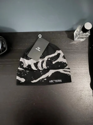 Svart och grå mössa från Arc'teryx - Snygg svart mössa från Arc'teryx med grått abstrakt mönster och broderad logga framtill. Perfekt för kalla dagar och streetwear-stil. Tunn och smidig modell som passar under jackhuvan. Helt ny.