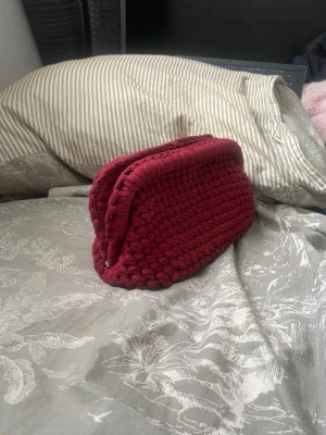 Handvirkad vinröd necessär - Säljer en handvirkad necessär i vinrött garn. Väskan har en mjuk, rektangulär form och är perfekt för att förvara smink eller småprylar. Den är gjord i ett tjockt, stickat material som ger en mysig känsla och unik look.