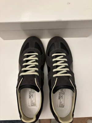 Maison Margiela  - Snygga Maison Margiela skor i mörkgrå mocka med svarta och grå detaljer. Skorna har vita snören och en klassisk gummisula i ljusbrunt. Ikonisk design med stilrena linjer och diskret branding på sulan. Perfekta för dig som gillar minimalistisk och trendig stil. Köpt i NK för 5999 kr. Priser kan diskuteras (inte under 4000) 