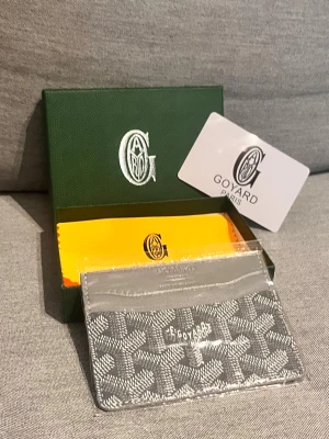 Grå Kortfodral från G - Snyggt grått kortfodral från G med det klassiska mönstret och vit logga framtill. Tillverkad i canvas och läder, perfekt för kort och ID. Kommer med originalask och certifikat. En stilren accessoar för dig som gillar exklusiva detaljer.