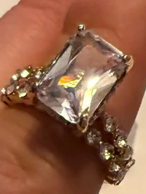 Guldfärgad ring med stor rektangulär sten - Säljer en guldfärgad ring med en stor, rektangulär klar sten i mitten och mindre runda stenar längs bandet. Ringen har en glittrig och lyxig känsla, perfekt för dig som gillar statement-smycken. Materialet ser ut att vara metall med glas- eller kristallstenar. St 16