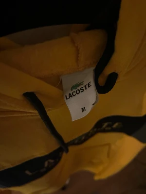 Gul hoodie från Lacoste - Gul hoodie från Lacoste med svart snörning i huvan och svart detalj över bröstet där Lacoste-loggan syns. Tillverkad i mjukt material som känns skönt mot huden. Perfekt för dig som vill sticka ut med en färgglad och sportig stil.