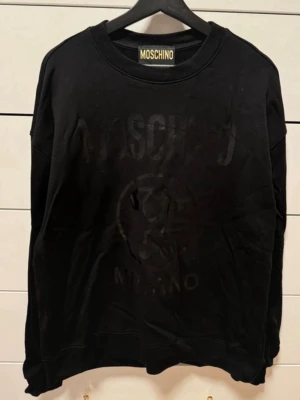 Svart sweatshirt från Moschino - Cool svart sweatshirt från Moschino med ton-i-ton logga och tryck framtill. Tröjan har rund halsringning och avslappnad passform. Perfekt för en trendig och avslappnad stil. Materialet känns mjukt och bekvämt mot huden.