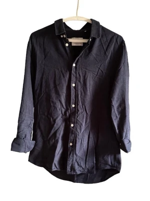 Mörkblå skjorta - Stilren mörkblå skjorta med klassisk krage och vita knappar framtill. Skjortan har lång ärm och är tillverkad i mjuk bomull, perfekt för en clean och enkel look.