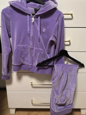 Lila velourset från Juicy Couture - Mysigt loungewear-set i lila velour från Juicy Couture. Setet består av en hoodie med dragkedja, fickor och broderad logga på bröstet samt matchande byxor med resår i midjan och fickor. Perfekt för chill dagar hemma. Hoddie i storlek XS & byxor i storlek M.