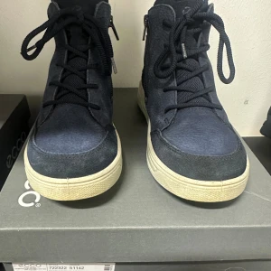 Mörkblå höga sneakers från Ecco - Snygga mörkblå höga sneakers från Ecco med vita sulor och svarta skosnören. Skorna har dragkedja på sidan och är tillverkade i en mix av textil och läder. Perfekta för dig som gillar stilrena och bekväma skor med sportig känsla.