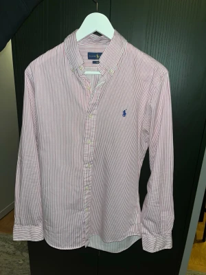 Rödvit randig skjorta från Ralph Lauren - Klassisk skjorta från Ralph Lauren med tunna röda och vita ränder. Skjortan har lång ärm, knappar framtill och den ikoniska blå logotypen broderad på bröstet. Tillverkad i mjuk bomull och har en tidlös, normal passform.