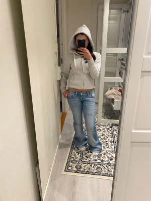 Ljusblå bootcut jeans med slitningar - Säljer ett par ljusblå bootcut jeans från Gina 14+ med slitningar på låren och snygga detaljer på bakfickorna. Jeansen har låg midja och klassisk femficksdesign. Perfekta för en avslappnad och trendig look.