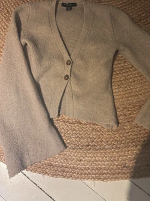 Beige kofta från Amisu med knappar - Mysig beige kofta från Amisu med två bruna knappar framtill. Koftan har v-ringning, lång ärm och är stickad i ett mjukt material. Perfekt att slänga över en t-shirt eller linne för en avslappnad look.