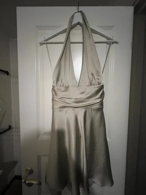Champagnefärgad halterneckklänning - Snygg kort klänning i champagnefärgat satinliknande material med halterneck och djup v-ringning. Knytband i ryggen för en extra detalj. Perfekt för dig som vill sticka ut med en elegant look. Oanvänd från zara