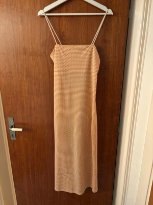 Lång orange/vit klänning med smala band - Säljer en lång klänning från Zara med smala axelband och ett diskret rutigt mönster i ljusa toner av orange och vitt. Klänningen har rak passform och är för varma dagar. Materialet känns lätt och luftigt, vilket gör den skön att bära. 
