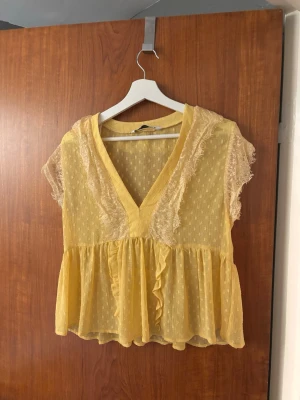Gul blus med spets och volang - Söt gul blus i transparent tyg med prickigt mönster, spetsdetaljer längs ärmar och axlar samt volang nertill. Djup v-ringning framtill och korta ärmar ger en luftig och somrig känsla. 