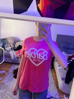 Rosa Barbie t-shirt med hjärta - Snygg rosa t-shirt från Barbie med ett stort vitt hjärta och texten 'Barbie Est 1959' på bröstet. T-shirten har rund halsringning och korta ärmar. Perfekt för dig som vill ha en lekfull och trendig look.