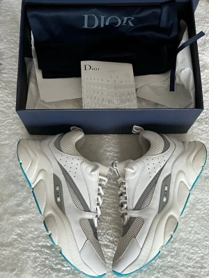 Vita chunky sneakers från Dior - Säljer ett par vita chunky sneakers från Dior med grå och silverdetaljer. Skorna har meshpaneler för extra ventilation, robust sula med turkosa inslag undertill och klassisk snörning. Perfekta för dig som gillar exklusiv streetwear och vill sticka ut.