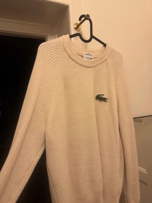 Beige stickad tröja från Lacoste - Säljer en beige stickad tröja från Lacoste med klassisk rund halsringning och det ikoniska gröna krokodilmärket på bröstet. Tröjan har lång ärm och ribbstickade muddar. Perfekt för dig som gillar stilrena och tidlösa plagg. Använd några få gånger. Som ny