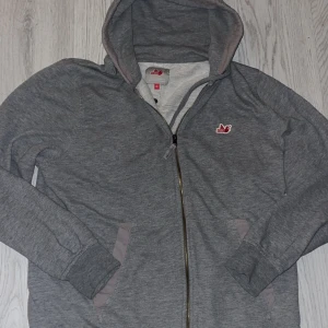 Grå zip hoodie från Northern Hooligans - Snygg grå hoodie med dragkedja från Northern Hooligans. Klassisk design med huva, två fickor fram och diskret logga i rött och vitt på bröstet. Mjuk insida och detaljer i ljusare grått vid fickor och huva. Perfekt för chill dagar eller streetwear-stil. 