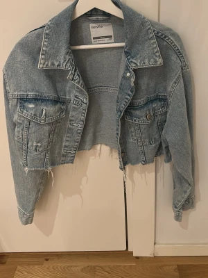 Cropped jeansjacka från Bershka - Trendigt croppad jeansjacka från Bershka i ljusblå denim. Jackan har råa, fransiga kanter och slitna detaljer på både ärmar och fickor. Klassisk krage, två bröstfickor med lock och knapp samt knappstängning framtill. Perfekt för en edgy streetstyle-look.