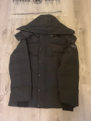 Svart dunjacka - Snygg svart dunjacka med huva och klassisk logotyp på ärmen. Jackan har knäppning med både dragkedja och tryckknappar samt quiltad design för extra värme. Perfekt för kalla vinterdagar.