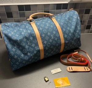 Louis Vuitton Keepall Monogram Denim - Stilren resväska från Louis Vuitton i blått denimtyg med klassiskt monogrammönster och ljusbruna läderdetaljer. Väskan har avtagbar axelrem, handtag och guldiga metalldetaljer. Perfekt för weekendresor och har en lyxig känsla.