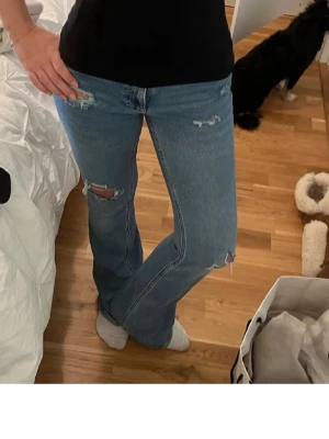 Blå slitna bootcut jeans - Säljer ett par blå bootcut jeans med slitningar och hål på låret och knät. Jeansen har klassisk femficksdesign och är i ett mjukt denimtyg. Perfekta för en avslappnad och trendig look.