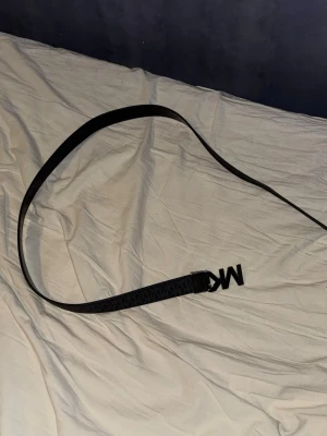 Svart Michael Kors bälte med MK-spänne - Snyggt svart bälte från Michael Kors med ett stilrent MK-spänne i metall. Bältet har diskret mönster längs hela remmen och passar perfekt till jeans eller kostymbyxor. Enkelt att justera för bästa passform.