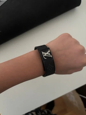 Louis Vuitton svart läderarmband - Snyggt svart armband från Louis Vuitton i läder med det klassiska LV-märket i silverfärgad metall. Armbandet har präglade monogramdetaljer längs hela bandet och en stilren, modern look som passar perfekt till din outfit.