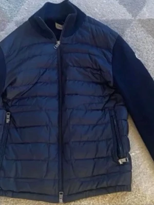 Svart dunjacka från Moncler - Snygg svart dunjacka från Moncler med quiltad front och rygg samt stickade ärmar. Jackan har dragkedja framtill, två fickor med dragkedja och Moncler-logga på ena ärmen. Perfekt för kyliga dagar och riktigt stilren look. Äkta jacka, Skicket är ungefär 9/10