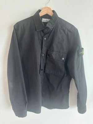 Svart overshirt från Stone Island - Stilren svart overshirt från Stone Island med klassisk krage, knappstängning och en bröstficka med lock. Ikonisk Stone Island-patch på vänster ärm. Tillverkad i ett slitstarkt bomullsmaterial som ger en clean och modern look.