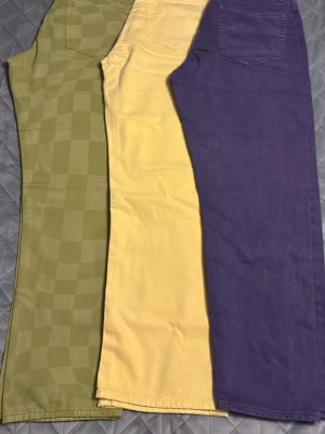 Tre färgglada raka jeans - Säljer tre par raka jeans i olika färger: ett par lila, ett par ljusgula och ett par ljusgröna med rutigt mönster. Alla har klassisk femficksdesign och hög midja. Perfekta för dig som vill sticka ut med färg och mönster i din outfit.