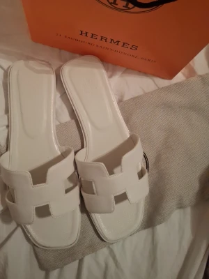 Vita  Oran sandaler - Säljer ett par vita Oran sandaler i äkta läder med den ikoniska H designen över foten. Sandalerna har platt sula och stilren, fyrkantig tå. Perfekta för en trendig och lyxig look. 