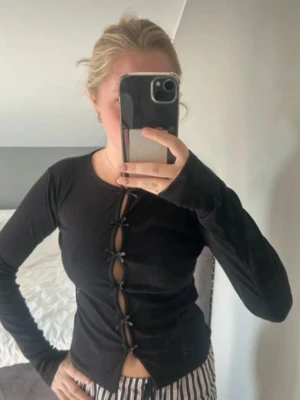Svart långärmad topp med knytning - Trendig svart långärmad topp med flera knytningar framtill som ger en cool cut-out-effekt. Rund halsringning och figurnära passform. Perfekt för dig som gillar en edgy och modern stil.