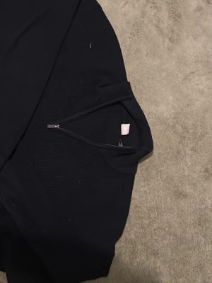 Svart ribbad tröja med halv dragkedja - Svart långärmad tröja med ribbad struktur och halv dragkedja vid halsen. Tröjan har hög krage och är tillverkad i ett mjukt, stretchigt material. Perfekt för en stilren och enkel look.