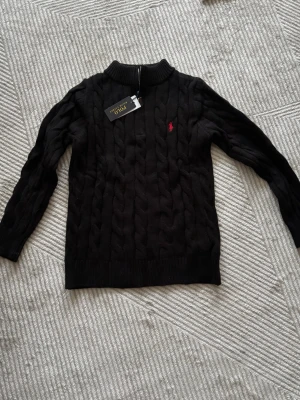 Svart kabelstickad tröja Polo Ralph Lauren - Svart kabelstickad tröja från Polo Ralph Lauren med klassisk röd logga broderad på bröstet. Tröjan har rund halsringning, ribbstickade muddar och är tillverkad i mjukt material. Perfekt för en stilren och tidlös look.