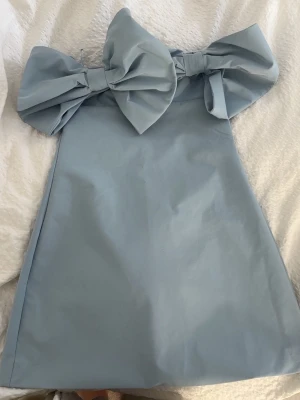 Ljusblå axelbandslös klänning med rosett - Supersöt ljusblå axelbandslös klänning med två stora rosetter framtill. Klänningen har en rak, kort siluett och är perfekt för dig som vill sticka ut. Materialet känns slätt och lätt, vilket ger en fräsch känsla. 