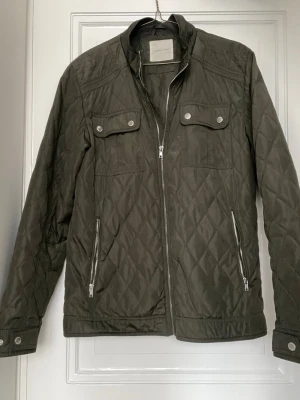 Quilted Jacks Selected Homme - Snygg olivgrön quiltad vindjacka från Selected Homme med dragkedja framtill, två bröstfickor med tryckknappar och två sidofickor med dragkedja. Jackan har ståkrage och detaljerade sömmar för en modern look. Perfekt för blåsiga dagar.