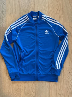 Blå Adidas Originals träningsjacka - Klassisk blå träningsjacka från Adidas Originals med vita ränder längs ärmarna och vit logga på bröstet. Jackan har dragkedja framtill, ribbade muddar och sidofickor. Tillverkad i ett glansigt, sportigt material som passar perfekt till träning eller streetwear. Stl S / Junior ca 14-16 år. Längd: 61 cm Bredd: 46/47 cm